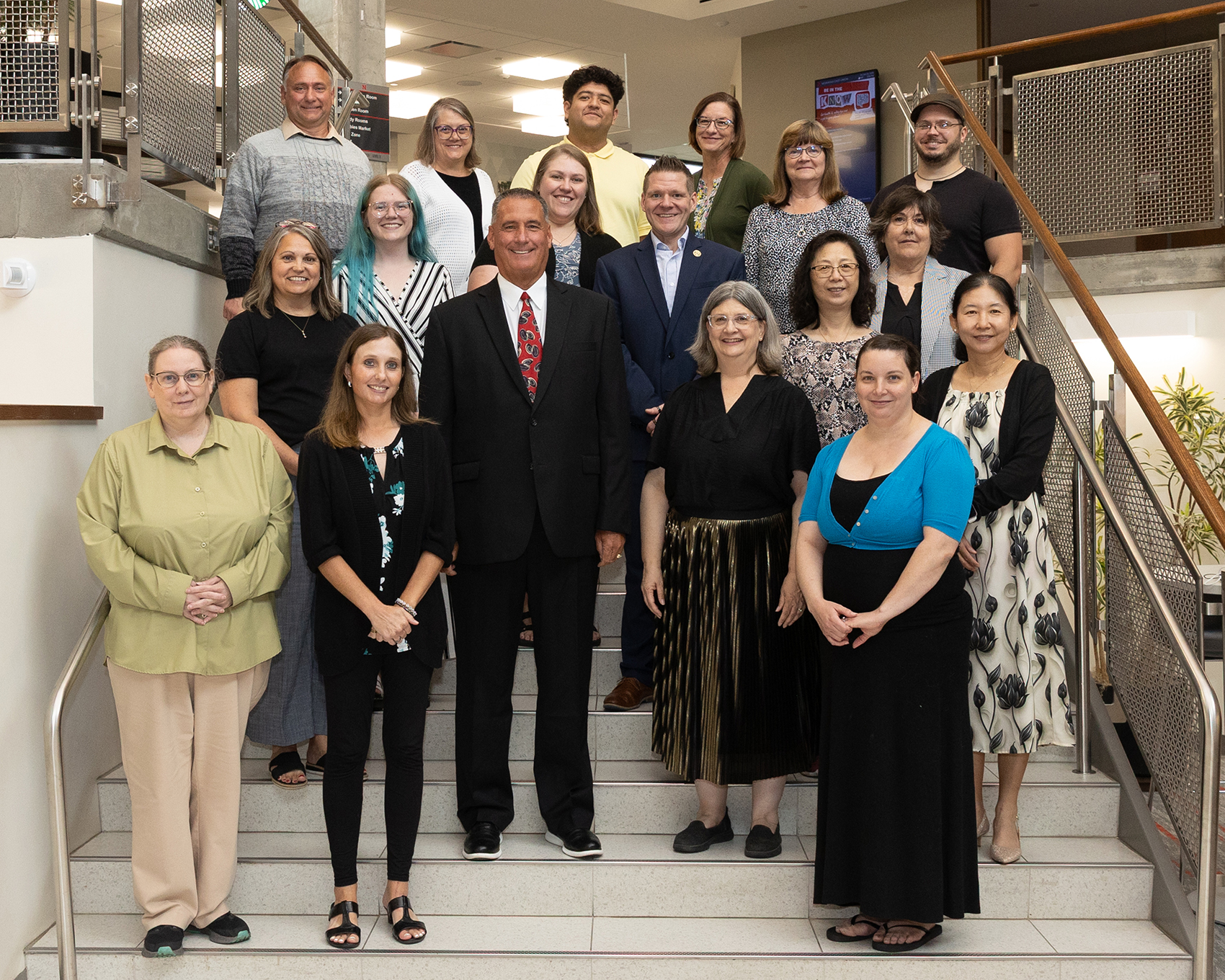Group photo of 18 University of Nebraska High School staff members standing on an indoor staircase, smiling and facing the camera.